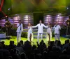 A group of performers in white outfits sings on stage accompanied by musicians while an audience watches