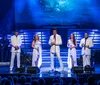A group of performers in white outfits sings on stage accompanied by musicians while an audience watches