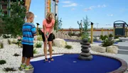 A woman is playing mini-golf with a boy on a course with blue artificial turf, surrounded by decorative rocks and plants.