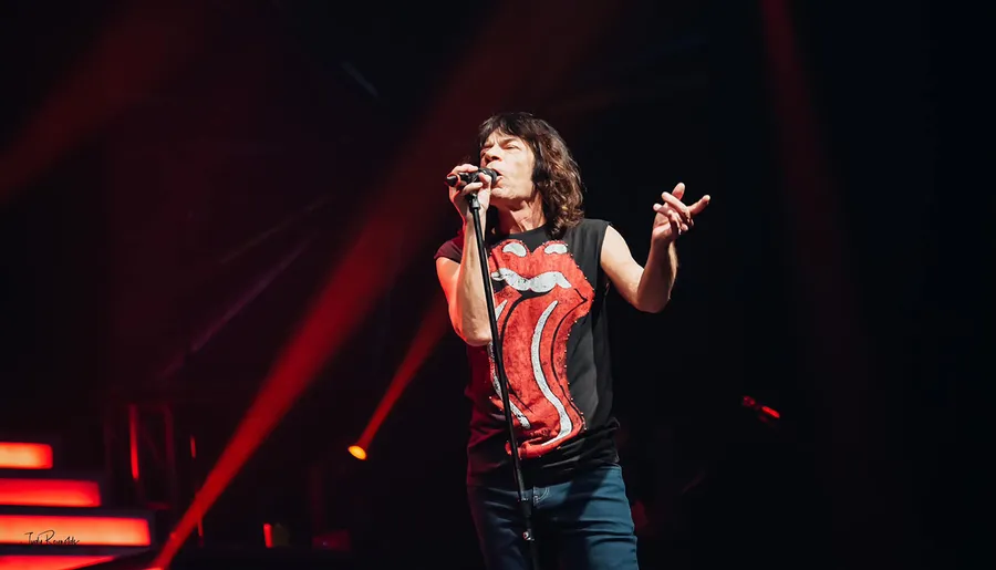 A person sings passionately on stage, wearing a Rolling Stones T-shirt, with red spotlights illuminating the scene.