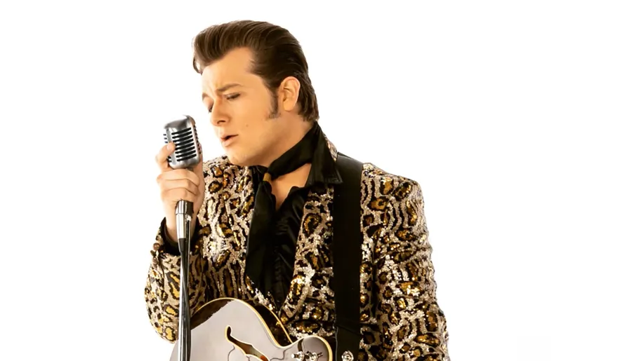 A person dressed in a leopard-print jacket sings into a vintage microphone while holding a guitar.