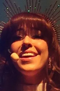 A person wearing a sparkling dress and a headpiece is smiling broadly with their arms outstretched on stage under a spotlight.