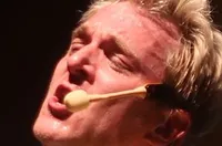 A person with spiked hair and a glittery gold vest and bow tie appears to be performing on stage, possibly singing or acting with an expressive facial expression.
