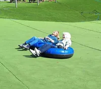 Two people are smiling and enjoying a ride down a green artificial slope on a large blue inflatable tube.