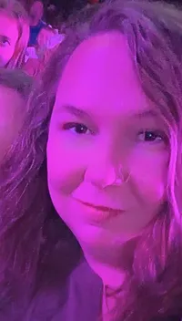 The image shows two people smiling and posing for a selfie in a purple-lit environment, with a group of people seated in the background.