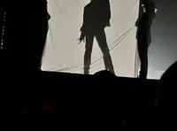 A silhouetted figure, wearing a hat, poses dramatically behind a backlit sheet held up by two people.