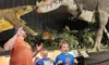 Three people pose humorously with expressions of surprise or fear in front of a large dinosaur exhibit.