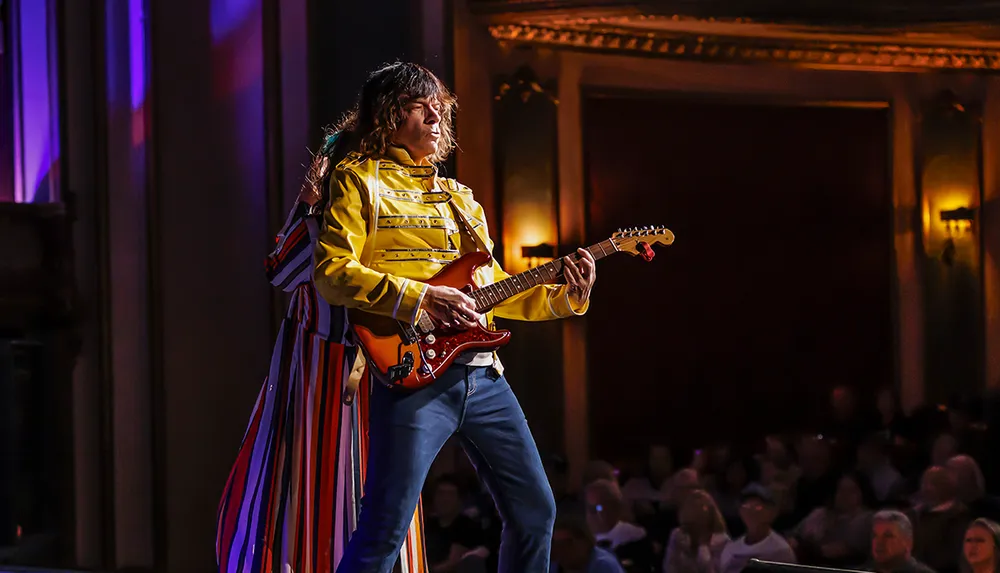 A musician in a yellow jacket plays an electric guitar on stage in front of an audience
