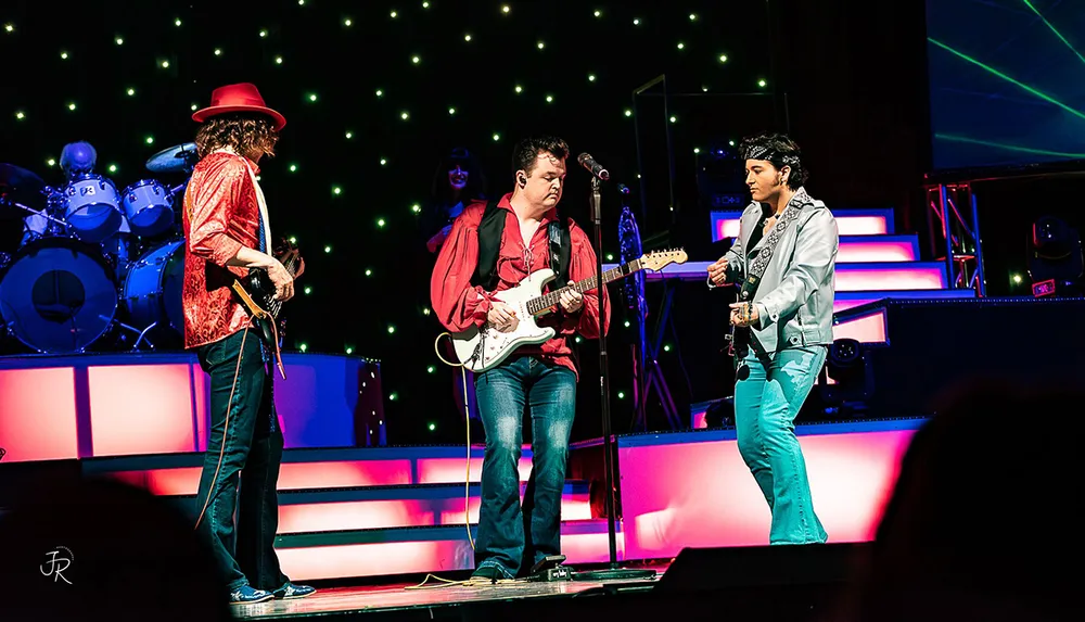 Three musicians are performing on stage with guitars vibrant lighting and a drummer in the background