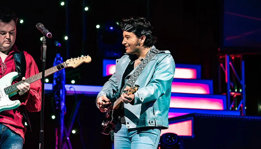 Two musicians are playing electric guitars on stage with colorful lights in the background