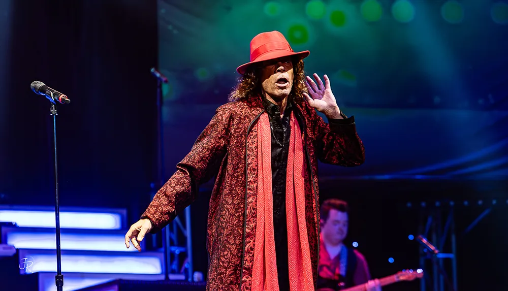 A performer dressed in a red and black patterned outfit and hat is on stage under colorful lights gesturing expressively