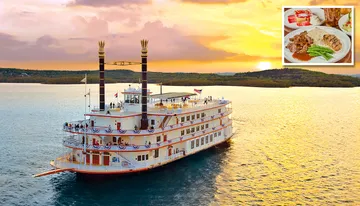 A paddle steamboat adorned with flags and bunting sails on a calm body of water during a vibrant sunset.