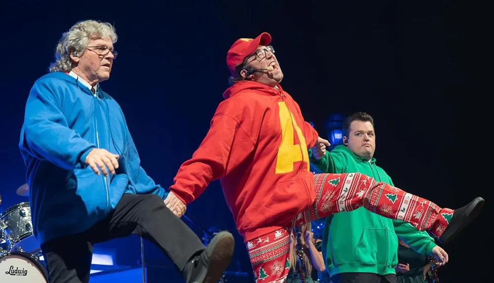 Three people are performing on stage energetically dancing and wearing colorful hoodies and festive pants