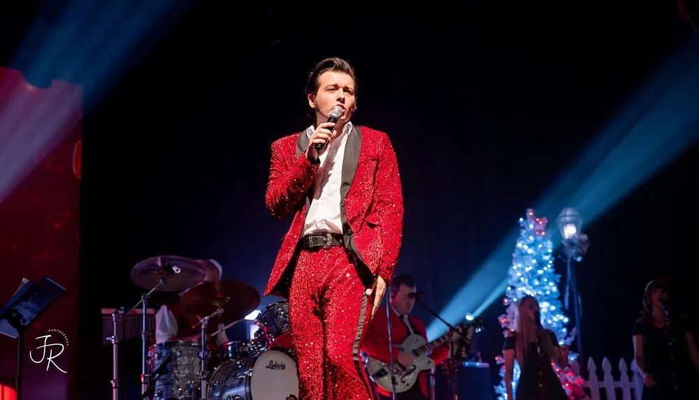 A performer in a sparkling red suit sings on stage accompanied by a band with a lit Christmas tree in the background