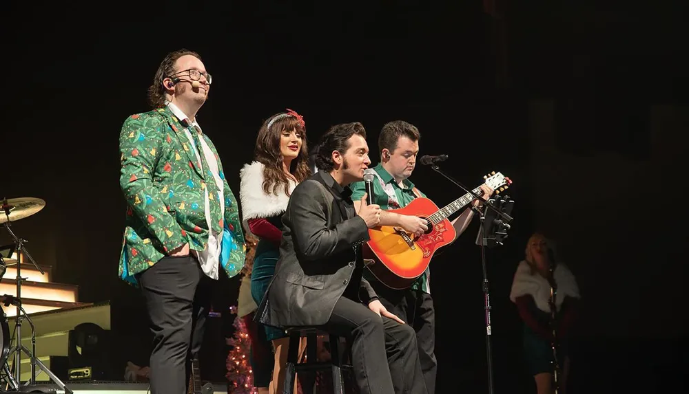 A group of musicians performs on stage with one person playing a guitar while another sings into a microphone and two others in festive attire