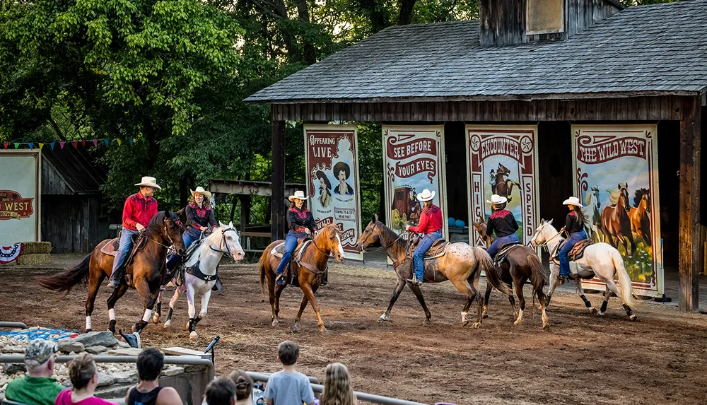 A group of people dressed in cowboy attire are riding horses in front of a rustic building with Wild West-themed posters while a small audience watches