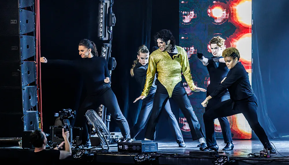 A performer in a gold shirt and black pants is dancing on stage with a group of dancers in black attire against a colorful backdrop while a camera operator captures the scene from the wings