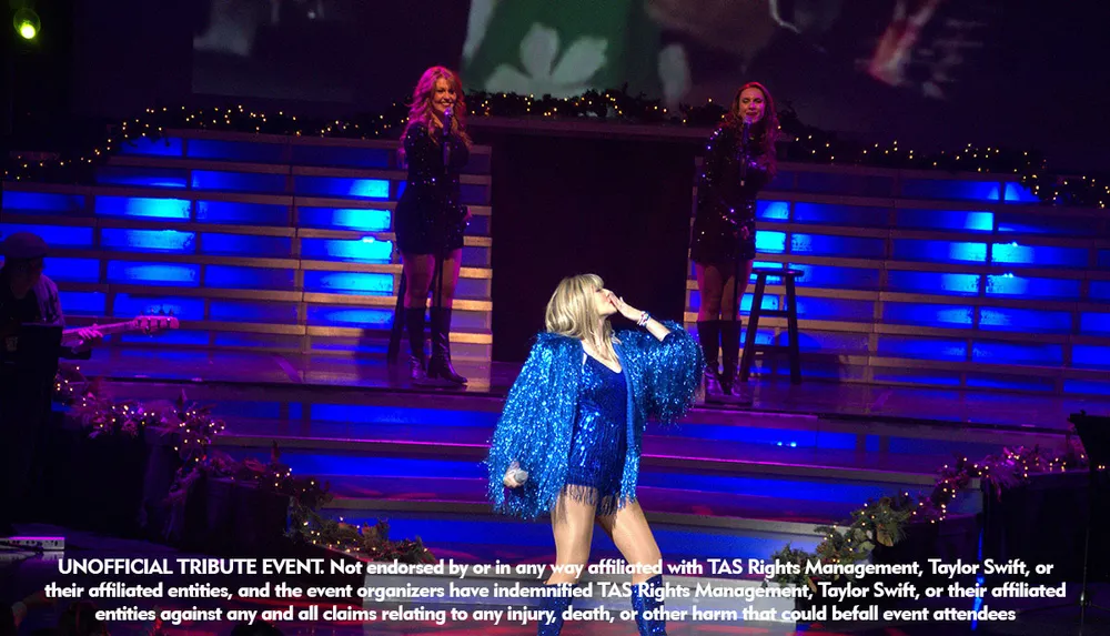 A singer in a blue outfit performs energetically on stage with two backup singers amidst festive decorations and blue lighting