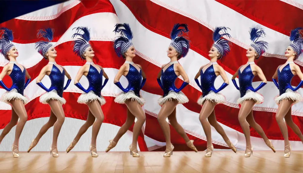 A line of dancers in sparkling blue costumes and feathered headpieces perform in front of an American flag backdrop