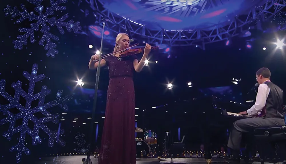 A woman is playing the violin on stage next to a man at a grand piano with a backdrop of snowflake decorations and dramatic lighting