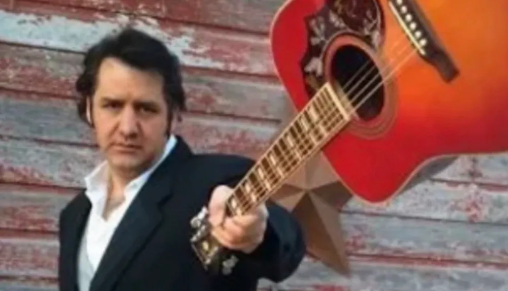 A person in a suit is holding a guitar in front of a weathered wooden wall