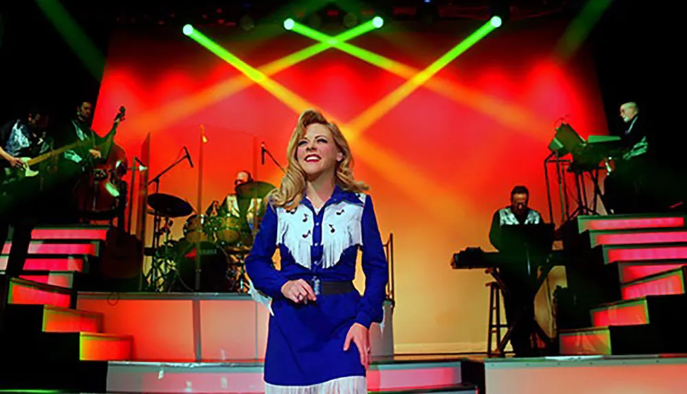 A performer in a blue and white outfit sings on a stage with a band in the background illuminated by colorful stage lights