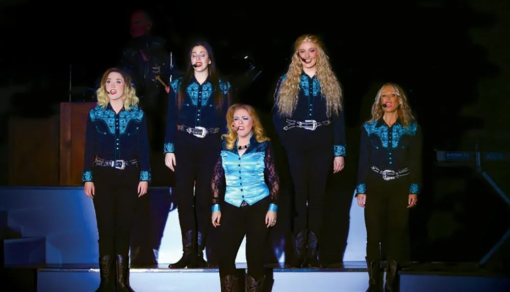 Five women wearing matching blue and black outfits with microphones perform on stage