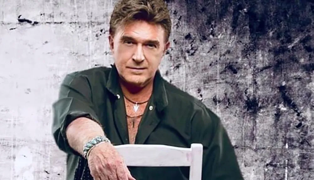 A man wearing a dark shirt and bracelets poses with his arm on the back of a chair against a textured background