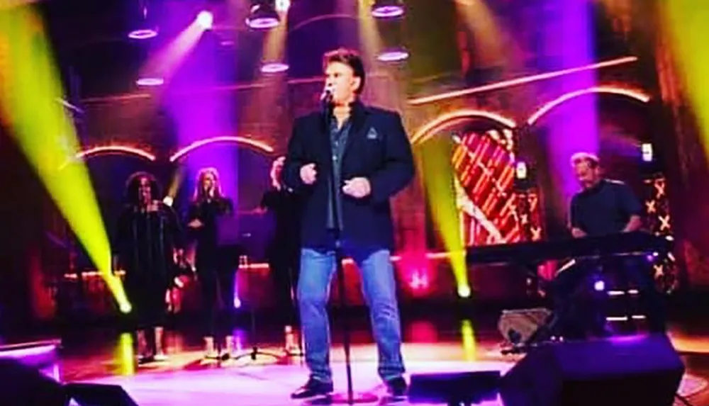 A performer is singing on stage with colorful lighting accompanied by backup singers and a keyboard player