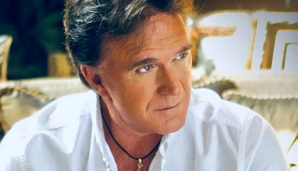 A man in a white shirt is looking off-camera with a thoughtful expression indoors
