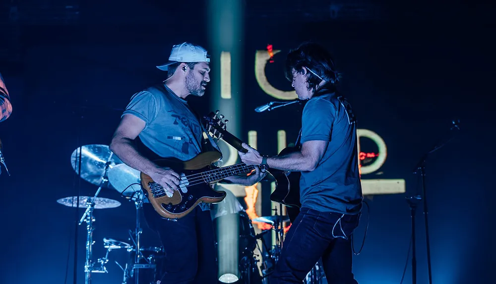 Two musicians are performing on stage one playing a bass guitar and the other a guitar with drum equipment visible in the background