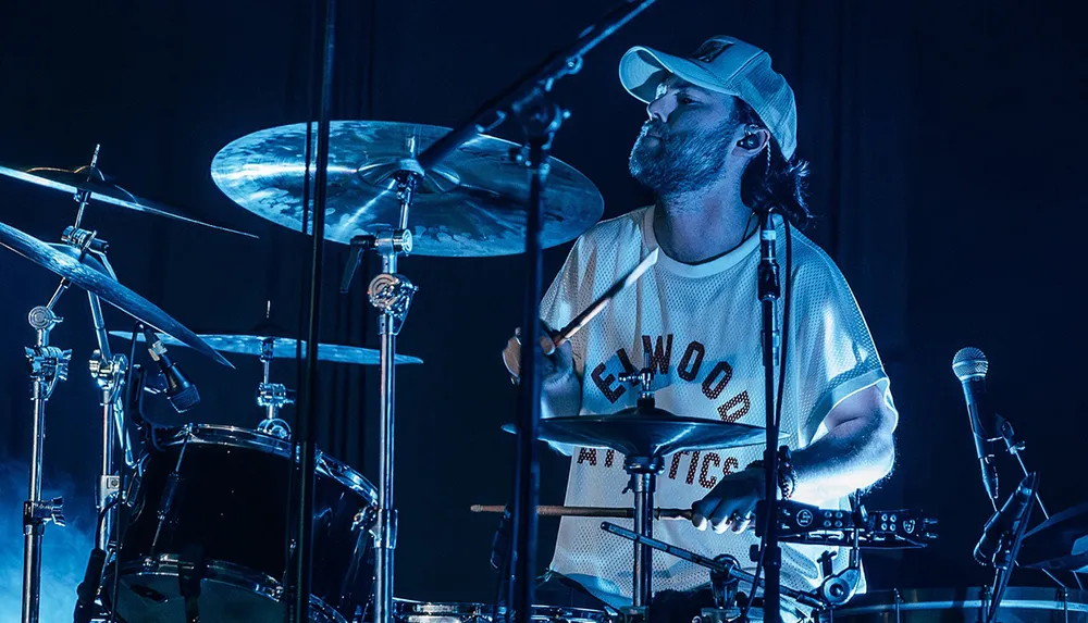 A drummer is energetically playing the drums on stage wearing a white cap and a shirt with ELWOOD ATHLETICS printed on it