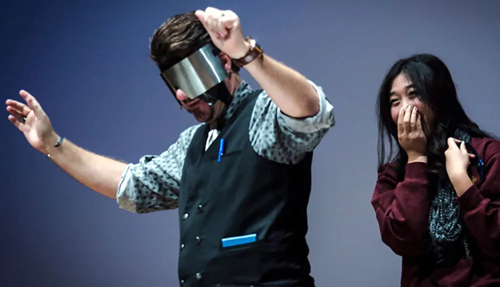 A man wearing a visor is performing on stage while a woman stands nearby smiling and holding her hand to her mouth