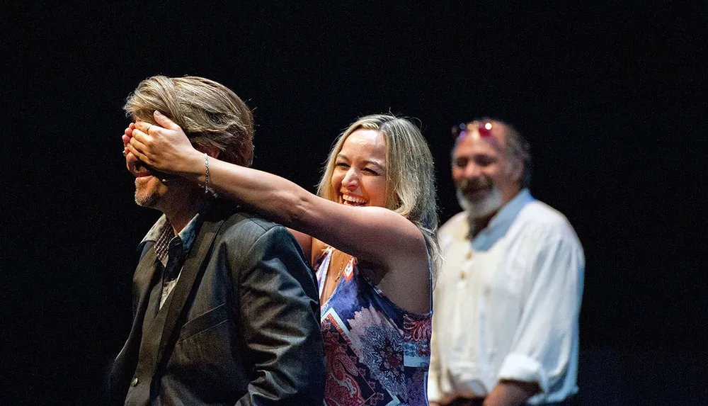 A woman playfully covers a mans eyes while another man watches and laughs in the background