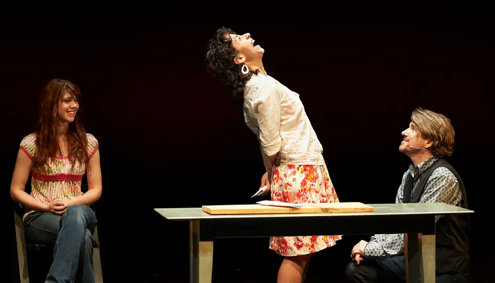 Three people are engaged in a theatrical performance with one person standing energetically between two seated individuals each displaying different expressions and postures