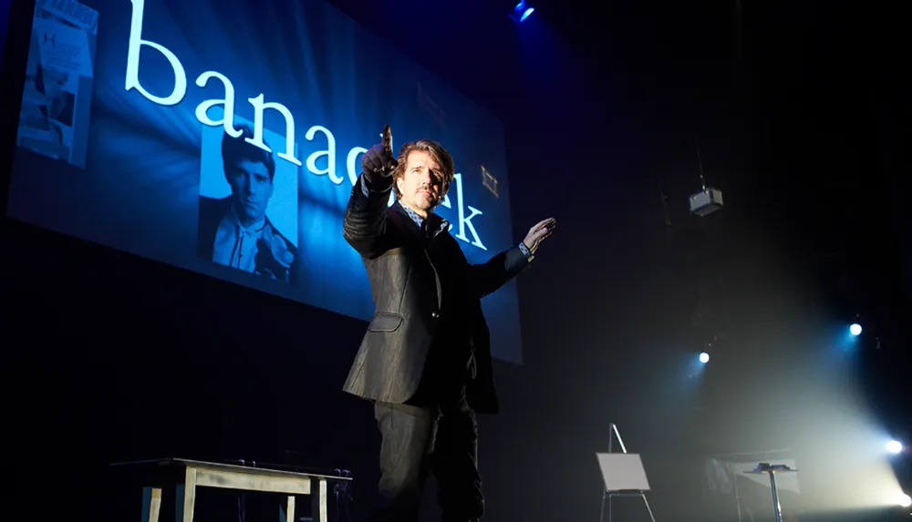 A man in a suit gestures theatrically on stage in front of a large screen displaying the text banachek