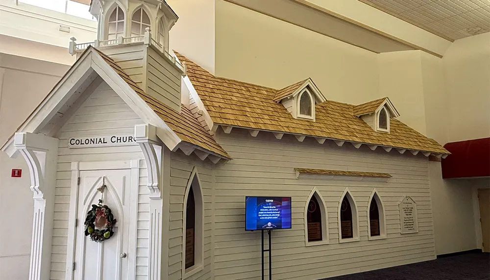 The image shows a small replica Colonial-style church with white siding arched windows a shingled roof and a digital screen displayed on the side