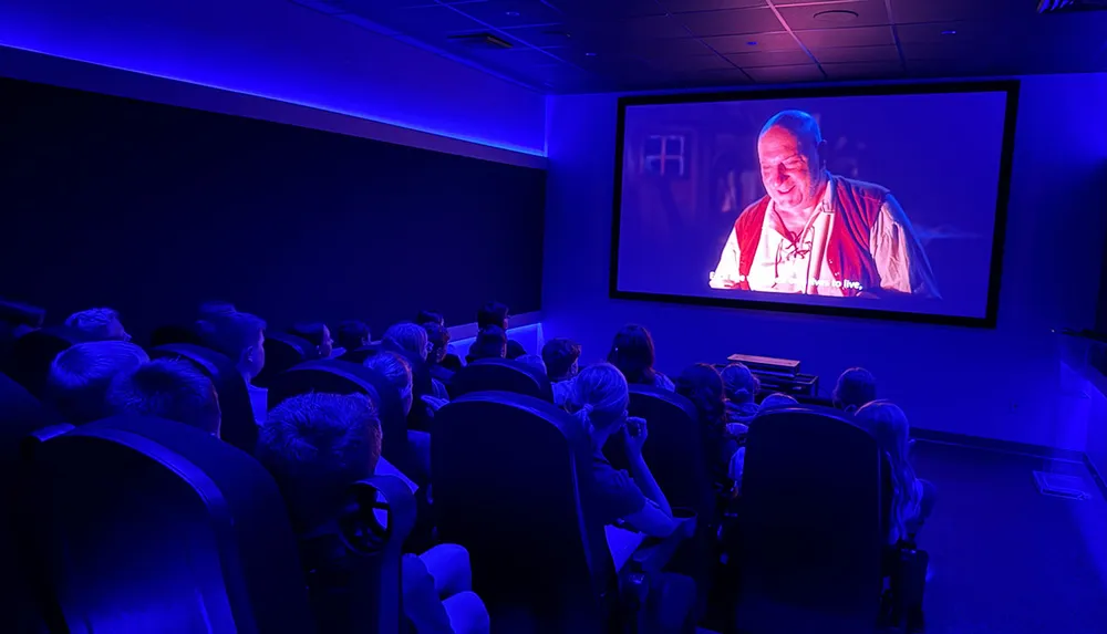 A group of people seated in a dimly lit room with a blue hue are watching a movie projected on a large screen