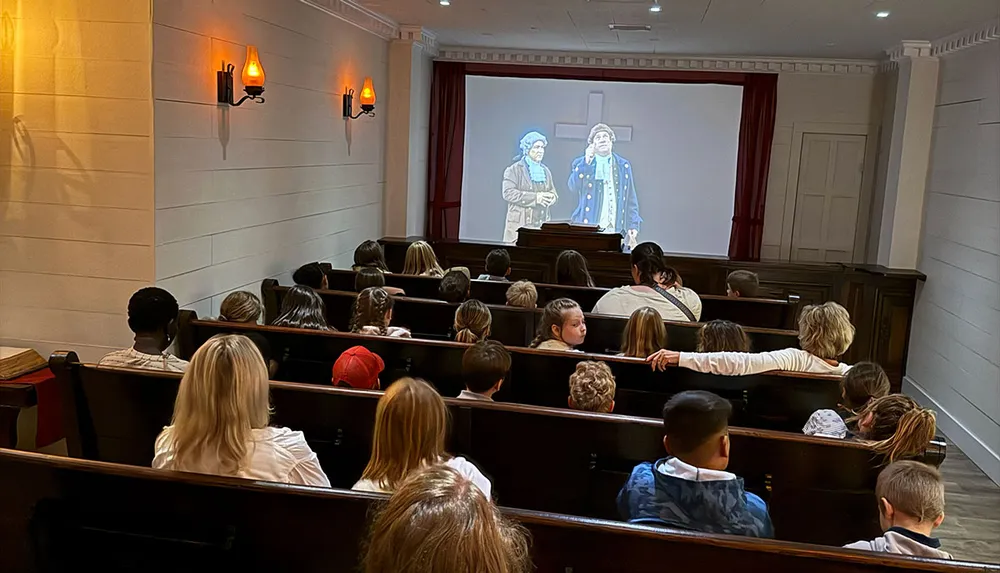 A group of people including children are seated in a dimly lit room watching a projected video of two historical figures