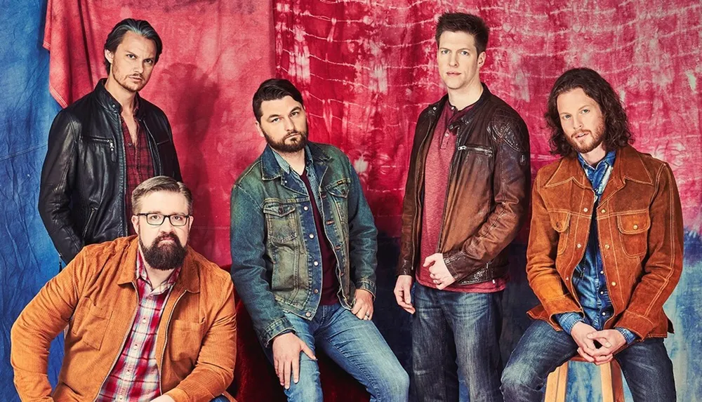 Five men in casual outfits are posing in front of a red and blue tie-dye background