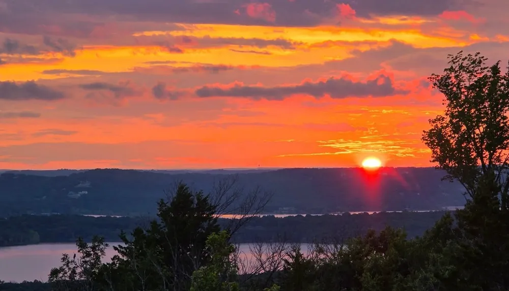 A vibrant sunset illuminates the sky with hues of orange and pink above a serene landscape featuring a lake and silhouetted trees