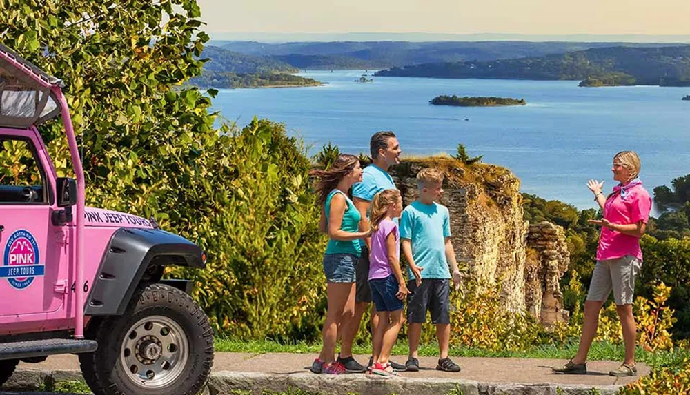 A tour guide talks to a family near a pink jeep with a scenic view of a lake and hills in the background