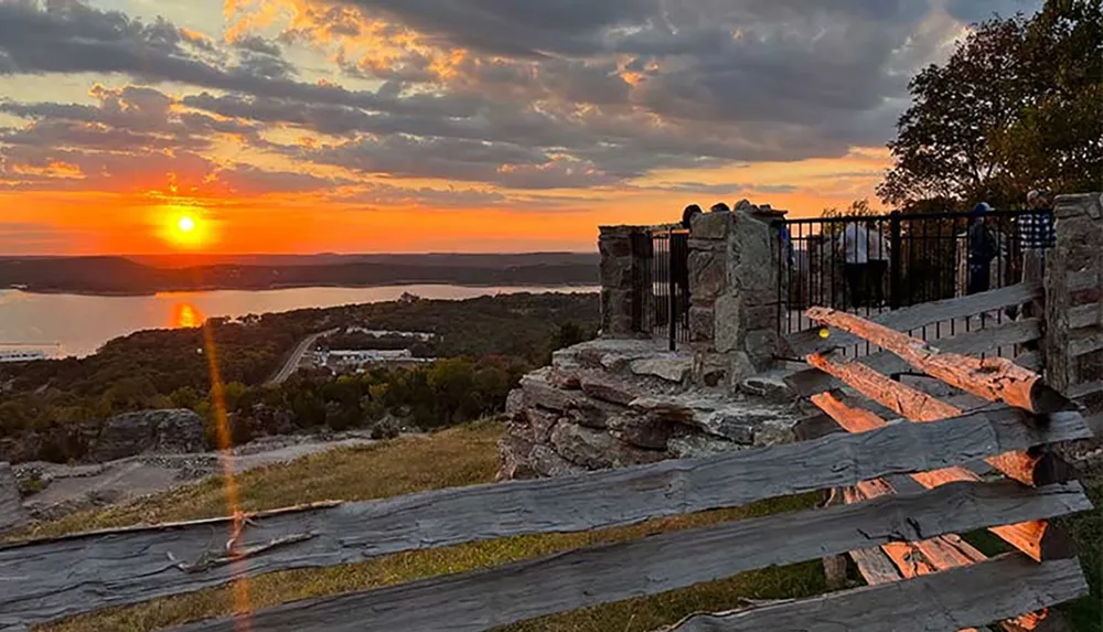 A scenic sunset view with the sun reflecting over a body of water and silhouetted people near a stone structure and wooden fence