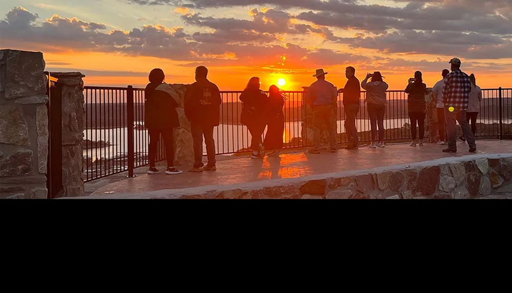 People are gathered at a viewpoint watching a vibrant sunset over a body of water with the sky filled with clouds