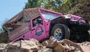 A pink Jeep is navigating a rocky off-road terrain amidst trees and rough slopes.