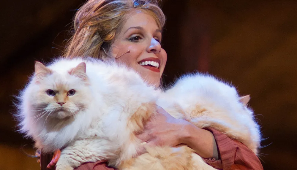 A smiling woman is holding a large fluffy white cat with an intense gaze