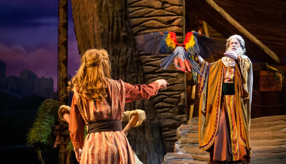A theater performer dressed in a colorful costume releases a brightly colored parrot towards another actor on stage set against a backdrop suggestive of a village or rustic setting at dusk