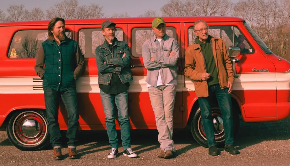 Four men are leaning against a vintage red and white van in a casual outdoor setting