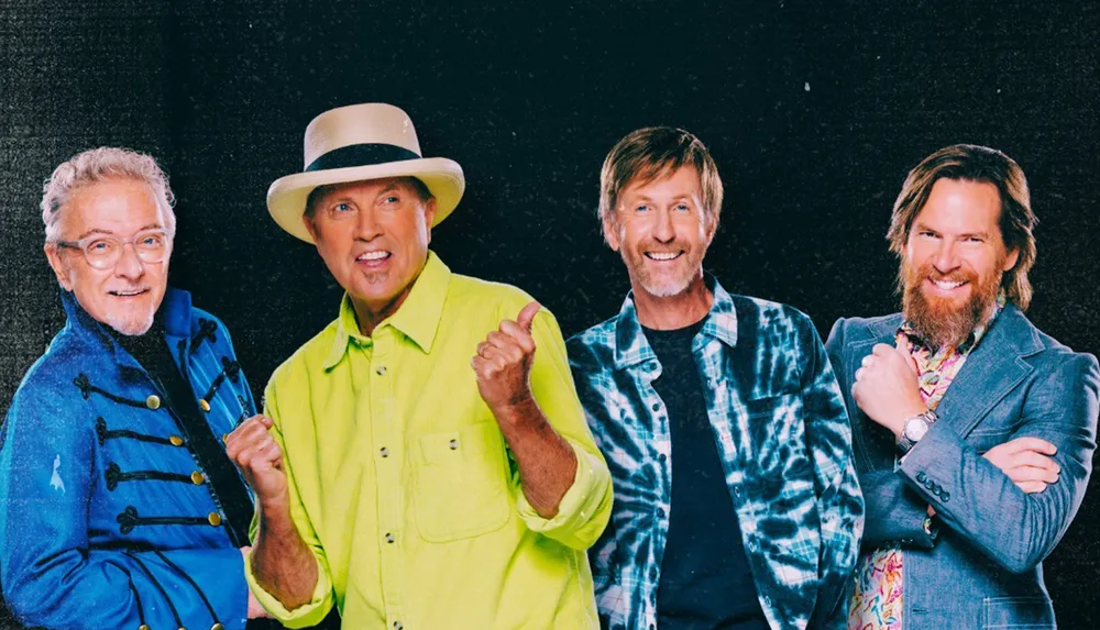 Four men are smiling and posing together against a dark background each dressed in colorful and distinctive clothing