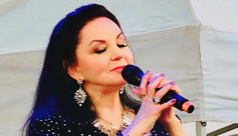 A woman with dark hair and elegant earrings is singing into a microphone with her eyes closed expressing emotion in her performance